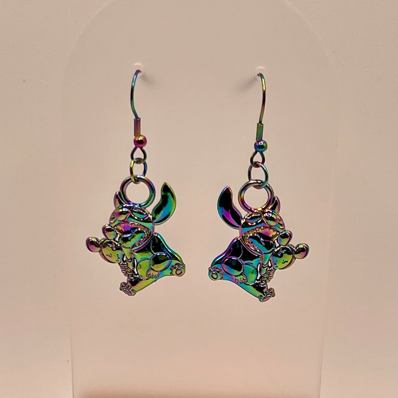 May include: A pair of iridescent silver earrings featuring a cartoon character with large ears and a mischievous expression. The character is holding a small object in its hand.