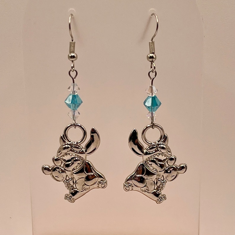 May include: Silver dangle earrings featuring a blue crystal charm and a silver Stitch character charm.