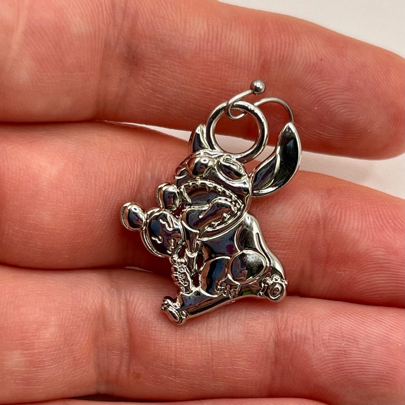 K&ouml;nnte beinhalten: Ein silberfarbener Anh&auml;nger mit der Figur Stitch aus dem Zeichentrickfilm Lilo & Stitch. Der Anh&auml;nger ist in sitzender Position und h&auml;lt ein kleines Objekt. Er hat eine Schlaufe zum Befestigen und ist sehr detailliert.