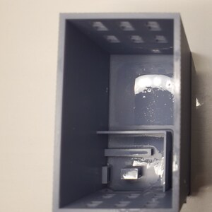 May include: A gray plastic box with a small, rectangular opening in the front. The box has a series of small holes on the top and bottom.