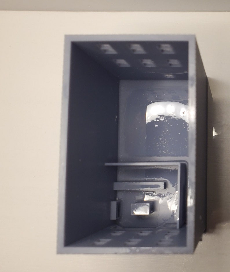 May include: A gray plastic box with a small, rectangular opening in the front. The box has a series of small holes on the top and bottom.