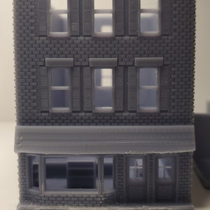 May include: A gray 3D printed model of a brick building with a storefront. The building has three floors with windows and shutters. The storefront has a large window and a door.
