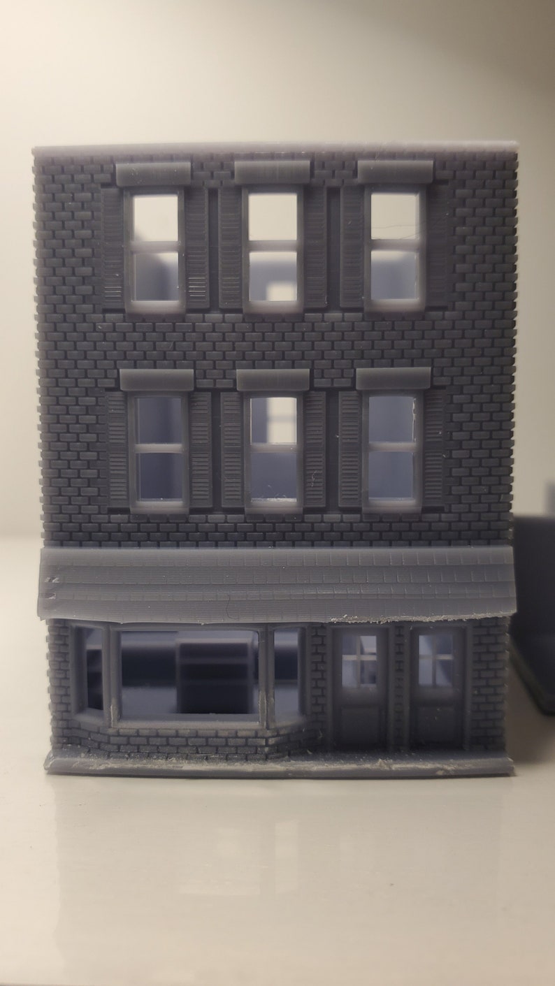 May include: A gray 3D printed model of a brick building with a storefront. The building has three floors with windows and shutters. The storefront has a large window and a door.