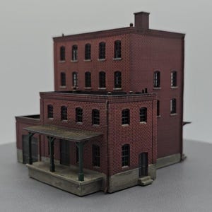 May include: A miniature brick building model with multiple stories, arched windows, and a covered porch. The model is primarily red brick with a dark roof and trim. The building has a chimney and a loading dock.