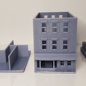 May include: A 3D printed model of a two-story brick building with a storefront. The model is disassembled into five pieces: the building itself, two side walls, a back wall, and a roof. The building has a gray color.