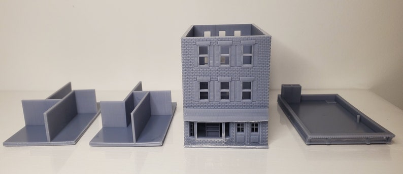 May include: A 3D printed model of a two-story brick building with a storefront. The model is disassembled into five pieces: the building itself, two side walls, a back wall, and a roof. The building has a gray color.