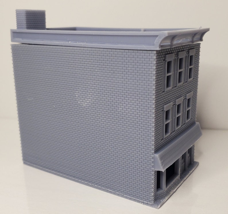 May include: A gray brick building model with a flat roof and a storefront awning. The building has three windows on each of the two upper floors.