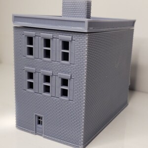 May include: A gray brick building model with a flat roof and a chimney. The building has three floors with windows on each floor. The windows have shutters. There is a door on the bottom floor.