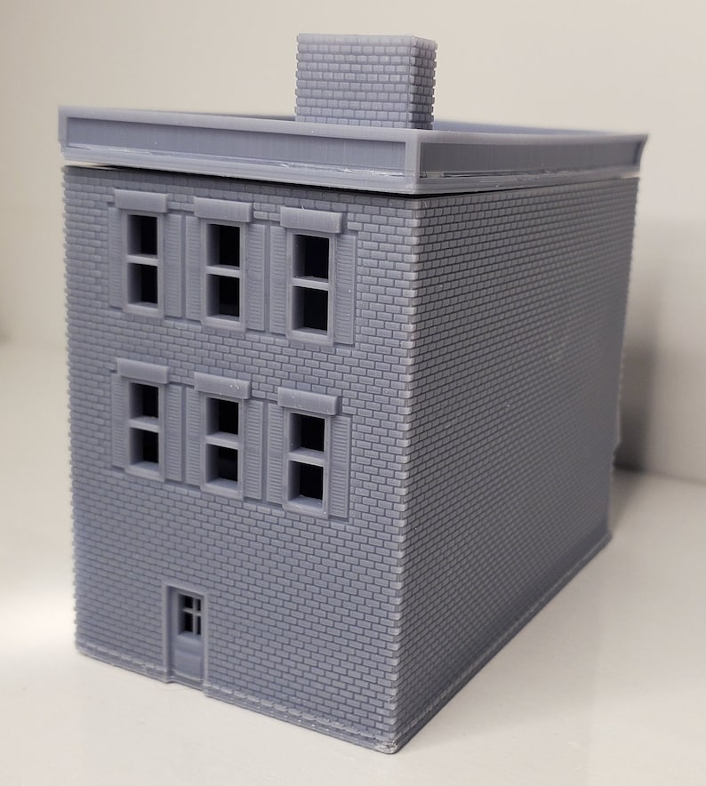 May include: A gray brick building model with a flat roof and a chimney. The building has three floors with windows on each floor. The windows have shutters. There is a door on the bottom floor.