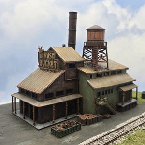 May include: A detailed model of a weathered industrial building, featuring a brick facade, corrugated metal roofing, and a prominent sign that reads "RUST BUCKET." The model includes a water tower, smokestack, and a loading dock, all set against a cloudy sky.
