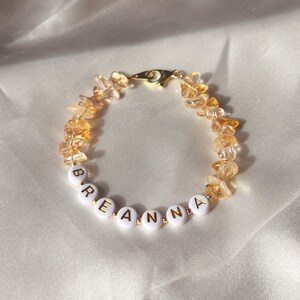 May include: A gold bracelet with a name spelled out in white beads. The name is "Breanna". The bracelet is made with small, clear, and amber-colored stones.
