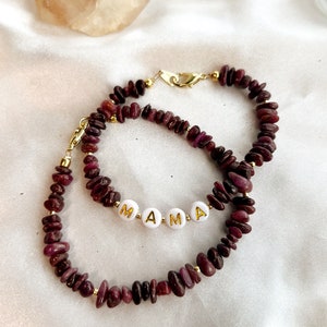 May include: Two bracelets made with small, dark red gemstone beads and gold accents. The bracelets are layered and the top bracelet has the word "MAMA" spelled out in white beads with gold lettering.
