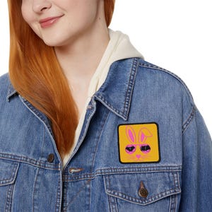 Könnte beinhalten: Eine blaue Jeansjacke mit einem quadratischen Aufnäher. Der Aufnäher zeigt ein Cartoon-Hasengesicht mit Sonnenbrille auf gelbem Grund. Die Jacke hat einen Knopfverschluss und wird über einem cremefarbenen Kapuzenpullover getragen.
