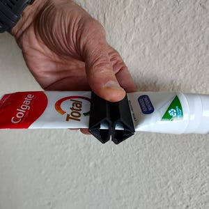 May include: A black plastic toothpaste tube squeezer is used to dispense toothpaste from a red and white Colgate Total tube. The tube is held horizontally, with the squeezer at the end.