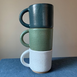 Puede incluir: Tres tazas de cerámica apiladas una encima de la otra. La taza superior es azul oscuro, la taza del medio es verde con motas blancas y la taza inferior es blanca con motas negras.