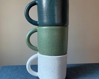 Peut inclure: Une pile verticale de trois tasses en céramique avec des anses arrondies. La tasse du haut est bleu foncé, celle du milieu est vert sauge et celle du bas est blanche. Chaque tasse présente une texture mouchetée sur toute la surface.