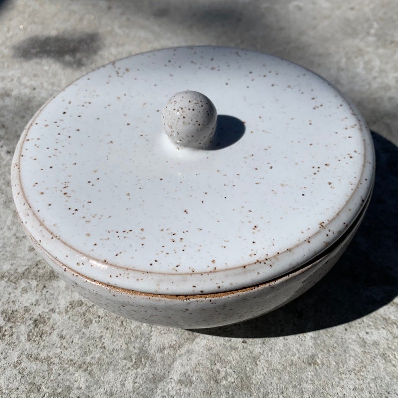 Peut inclure: Bol en c&eacute;ramique blanche avec un gla&ccedil;age mouchet&eacute; et un couvercle avec un bouton. Le bol et le couvercle ont une forme l&eacute;g&egrave;rement arrondie.
