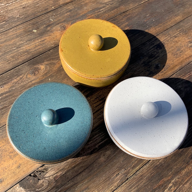 Peut inclure: Trois bols en c&eacute;ramique avec couvercle de diff&eacute;rentes couleurs. Les bols sont jaunes, bleus et blancs. Chaque bol a un bouton rond sur le couvercle.
