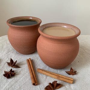 May include: Two terracotta clay cups, one filled with dark liquid and the other with a lighter beverage, sit side-by-side. Cinnamon sticks and star anise are scattered around the cups on a textured, off-white surface. The cups have a rustic, handmade appearance.