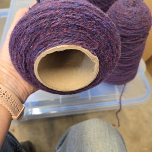 May include: Close-up of several large spools of yarn in shades of purple and red. The yarn is wound around a cardboard tube. The yarn is a textured, woven material. The spools are sitting on a clear plastic container.