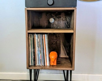 May include: A walnut wood record player stand with two shelves. The stand has black hairpin legs and is holding a record player and a stack of vinyl records. There is a small orange elephant figurine on the bottom shelf.
