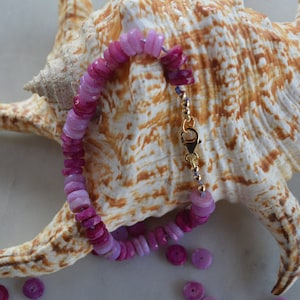 May include: A pink and purple gemstone bracelet with a gold clasp. The bracelet is made of round, flat beads and is lying on a seashell.