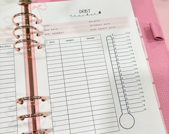 Puede incluir: Agenda rosa con anillos dorados y una hoja de seguimiento de deudas. La hoja tiene un gráfico de termómetro y secciones para fecha, pago y saldo. El texto "Debt Tracker" está escrito a mano. También se ve un letrero decorativo de purpurina "ezbudgets".