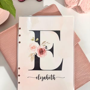 May include: A pink and white floral design with a black letter E and the name "elizabeth" written in cursive. The design is on a white background with a pink border. The design is on a sheet of paper with holes on the left side.