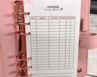 Può includere: Un'agenda rosa con finiture color oro rosa contiene un inserto bianco per il rilevamento delle spese. L'inserto ha un'intestazione che recita "expense tracker" e include colonne per data, spesa, categoria, importo, contanti e carta.