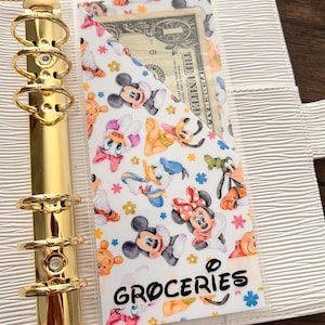 Puede incluir: Un bolsillo de plástico blanco y transparente para un organizador personal con un estampado de personajes de Disney que incluye a Mickey Mouse, Minnie Mouse, Donald Duck, Goofy y Pluto. El bolsillo tiene una etiqueta negra que dice "GROCERIES".