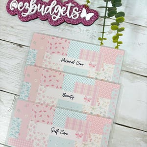 May include: Three clear plastic budget envelopes with a patchwork design in pink, white, and light blue. Each envelope is labeled with a different category: "Personal Care", "Beauty", and "Self Care". The top of the image has the text "ezbudgets".