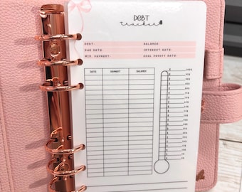 Può includere: Un'agenda rosa con elementi in oro rosa e un inserto "Debt Tracker". L'inserto ha un'intestazione rosa con campi per debito, data di scadenza e saldo. Un termometro è a destra, con una scala da 0 a 1000 €.