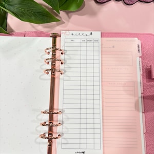 May include: A pink planner with a rose gold binder and a white bill tracker insert. The bill tracker has a grid for tracking bills, due dates, amounts, and if they are paid. The text "bills" is at the top of the insert.
