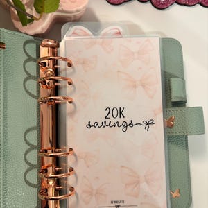 May include: A mint green planner with rose gold hardware and a cover featuring a bow pattern. The planner insert reads "20K savings". Decorative butterfly accents and the text "@ezbudgets" are also visible.
