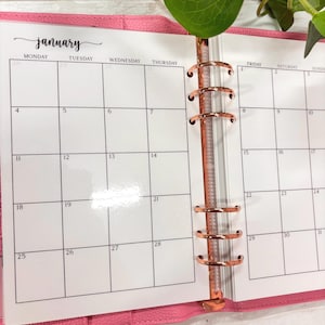 May include: A pink leather-textured planner open to a January calendar page. The white paper features black grid lines, days of the week, and dates. A metallic rose gold six-ring binder mechanism secures the pages within the pink cover.