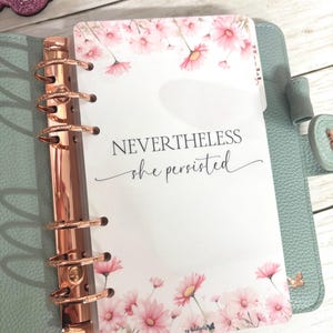 May include: A planner insert with pink daisy floral design and the text "NEVERTHELESS she persisted." The insert is inside a light blue planner with rose gold rings. The planner has a butterfly charm.