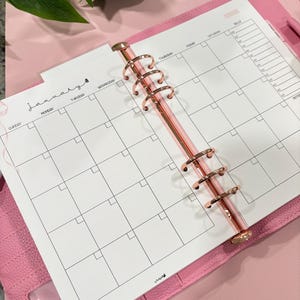May include: A pink planner with a rose gold binder, open to a January monthly calendar page. The calendar is white with black grid lines and text. The planner has a clear pocket and the text "yes budgets" is visible.