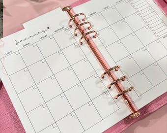 May include: A pink planner with a rose gold binder, open to a January monthly calendar page. The calendar is white with black grid lines and text. The planner has a clear pocket and the text "yes budgets" is visible.