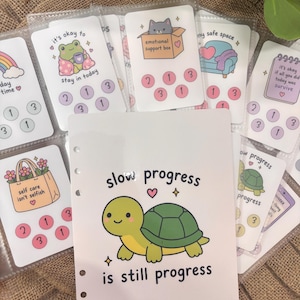 Può includere: Una collezione di schede di tracciamento in plastica bianca con illustrazioni colorate di una tartaruga, una rana, un gatto in una scatola e un divano. Le schede includono testi come slow progress is still progress e emotional support box.