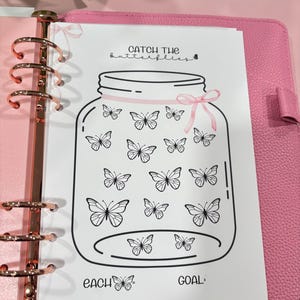 May include: A pink planner with rose gold rings holds a white page with a jar illustration. The jar is filled with butterfly drawings and the text "Catch the butterflies" and "Each Goal". A pink ribbon adorns the jar.