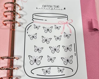 Può includere: Un'agenda rosa con anelli color oro rosa contiene una pagina bianca con un'illustrazione di un barattolo. Il barattolo è pieno di disegni di farfalle e il testo "Catch the butterflies" e "Each Goal". Un nastro rosa adorna il barattolo.