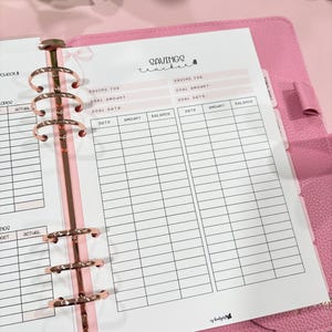 Peut inclure: Un agenda rose avec des anneaux en métal rose doré et une feuille "Savings Tracker". La feuille contient des sections pour les objectifs d'épargne, les dates, les montants et les soldes. L'agenda est ouvert, révélant les pages de suivi financier.