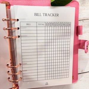 May include: A bright pink textured binder with rose gold rings holding a white paper insert titled BILL TRACKER. The insert features a grid layout with columns for bill names, total amounts, and individual months from January through December.
