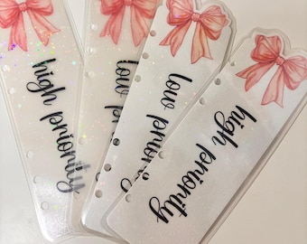 May include: Four clear planner dividers with a pink bow design and the words "high priority" and "low priority" in black script. The dividers have a glittery, sparkly effect and are pre-punched with holes for easy insertion into a planner.