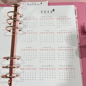 May include: A pink planner with a 2026 calendar, featuring monthly layouts. The planner has a rose gold spiral binding and dividers. The calendar pages display the days of the week and dates.