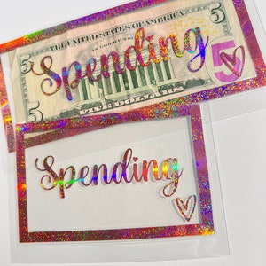 May include: Two rectangular clear plastic cash envelopes featuring a holographic glitter border. The top envelope contains a five-dollar bill. Both envelopes display the word Spending in holographic script lettering and a small heart outline.