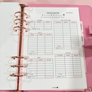 May include: Pink planner with rose gold rings, open to a "Paycheck Breakdown" sheet. The sheet has sections for income, bills, debt, and savings, with spaces for dates, amounts, and budget tracking. The planner is on a white surface.