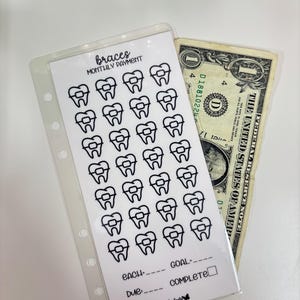 Puede incluir: Una funda de plástico transparente con una inserción blanca con la etiqueta "braces MONTHLY PAYMENT". La inserción presenta una cuadrícula de dientes con aparatos ortopédicos, un billete de dólar es parcialmente visible detrás de la funda. El texto "EACH", "GOAL" y "DUE" también es visible.