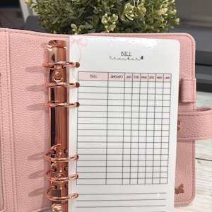 Peut inclure: Un classeur rose en similicuir avec une quincaillerie en or rose, ouvert pour révéler un insert de suivi des factures. L'insert est blanc avec une grille pour le suivi des factures, et les mots "Bill Tracker" sont écrits en haut.