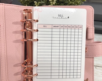 Può includere: Un raccoglitore rosa in similpelle con hardware color oro rosa, aperto per rivelare un inserto per il monitoraggio delle fatture. L'inserto è bianco con una griglia per il monitoraggio delle fatture e le parole "Bill Tracker" sono scritte in alto.